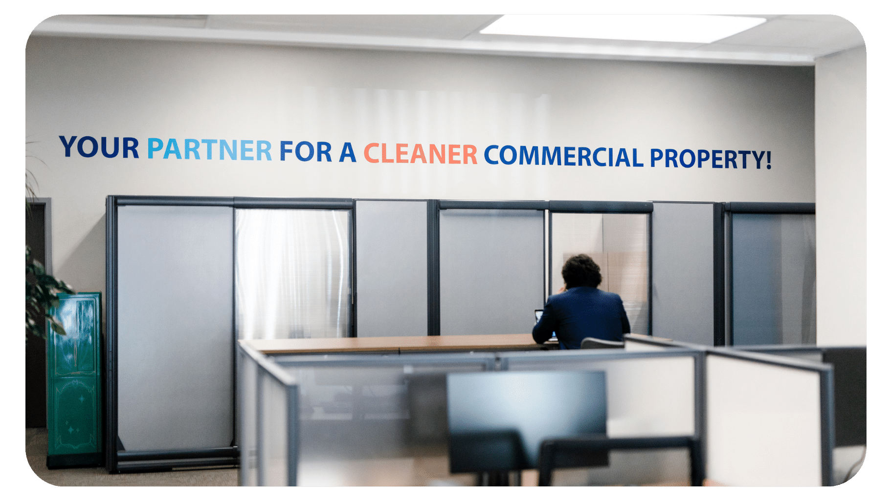 A person sits alone at a desk in a modern office with a wall sign reading, Your partner for a cleaner commercial property! in colorful letters.