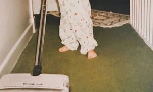 A child in pajamas stands barefoot on green carpet while pushing a vacuum sweeper down a hallway. The childs upper body is out of the frame.