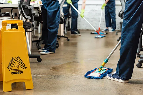 Several people from Impact Commercial Cleaning Services are mopping the floor in an office setting. A yellow caution sign warns of a wet floor, with desks, chairs, and cleaning equipment visible—a clear display of commercial cleaning in Canada.