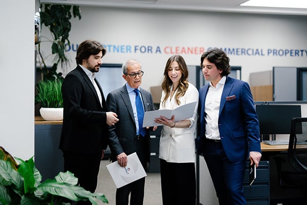 Four professionally dressed people stand together in an office, looking at documents and smiling. The workspace background includes desks, office plants, and a slogan on the wall—reflecting impact commercial cleaning services in Canada.