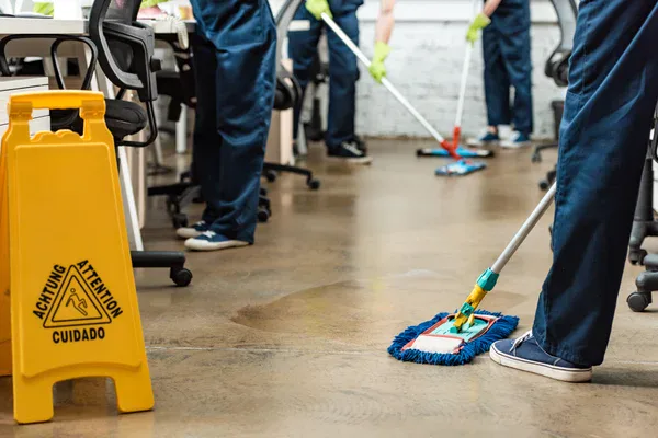 People in uniforms from impact commercial cleaning services clean an office floor with mops; a yellow caution wet floor sign is visible in the foreground. Desks and chairs are arranged in the background.