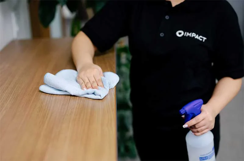 A person in a black IMPACT shirt wipes a wooden surface with a cloth while holding a spray bottle, representing impact commercial cleaning services in Canada.