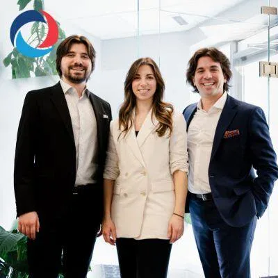 Three people, two men in suits and a woman in a white blazer, stand smiling together in a bright office space. A circular logo with blue and red is visible on the glass wall behind them, reflecting their role in commercial cleaning Canada.