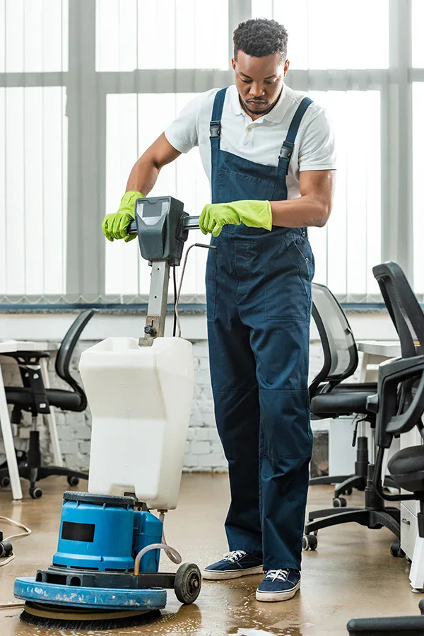 A man wearing overalls and green gloves uses a floor cleaning machine in an office with desks and chairs, demonstrating the professionalism of impact commercial cleaning services.