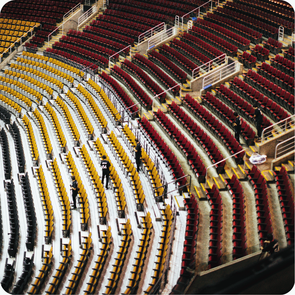 Rows of empty stadium seats in alternating red and yellow sections, with a few people scattered throughout the aisles. White railings divide the sections, and the angle is from above.