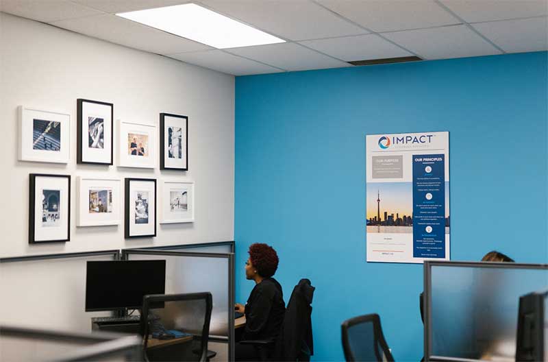 A person works at a desk in an office with blue and white walls, framed pictures, and an informational poster labeled Impact. Several empty workstations highlight the need for professional impact commercial cleaning services.