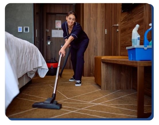 A woman in a housekeeping uniform vacuums the carpet in a hotel room, with a cleaning supplies caddy placed on a bench nearby.