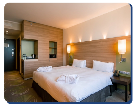 Modern hotel room with a large neatly made bed, two white robes, wooden headboard, wall lamps, a bedside table with a phone, and a kitchenette featuring a coffee maker and safe—kept pristine by commercial cleaning Canada.