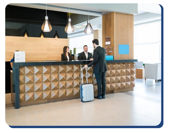 A man in a suit with a rolling suitcase stands at a modern hotel reception desk, speaking with two receptionists behind the counter. The lobby is bright with contemporary decor.