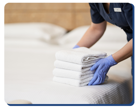 A person in blue gloves and a dark uniform from Impact Commercial Cleaning Services places a neatly folded stack of white towels on a made bed in a clean, tidy room.