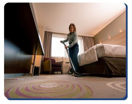A woman is vacuuming the carpet in a neatly arranged hotel room with a made bed, armchair, and large window with curtains.