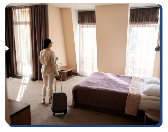 A person with a suitcase enters a brightly lit hotel room with large windows, a neatly made bed, and modern decor.