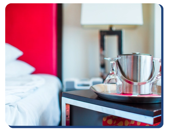 A hotel room with a neatly made bed, a bedside table holding an ice bucket, two upside-down glasses on a tray, and a modern lamp in the background.