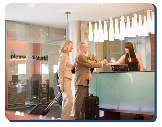 A man and woman with suitcases check in at a hotel reception desk, where a receptionist hands a key card to the man. The background shows a glass wall with “please disturb!” text.