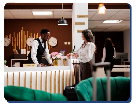 A hotel receptionist assists a woman with a suitcase at the front desk, with clocks and flowers in the background. Another person walks away in the lobby, which reflects the standards upheld by impact commercial cleaning services.