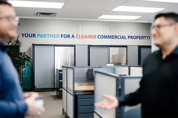 Two people talk in a modern office with cubicles. On the wall behind them, text reads: YOUR PARTNER FOR A CLEANER COMMERCIAL PROPERTY! Experience the difference with Impact Commercial Cleaning Services in Canada.
