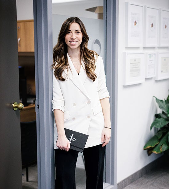 A woman with long brown hair, wearing a white blazer and black pants, stands smiling in an office doorway holding a black notebook. Framed certificates and a plant highlight the professional setting of impact commercial cleaning services.