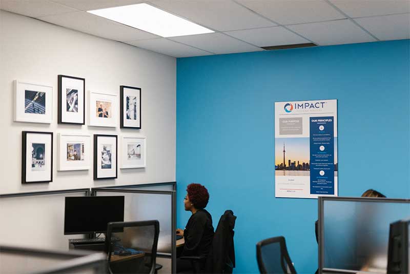 A person sits at a computer desk in a modern office with blue and white walls, framed photos, and an IMPACT informational poster promoting impact commercial cleaning services. Other empty workstations and chairs are visible.