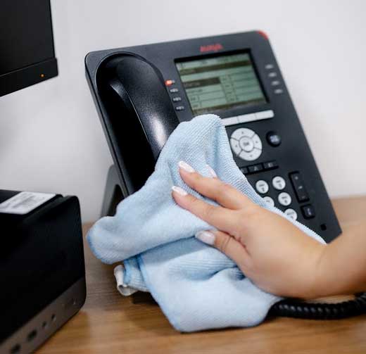 A person’s hand wipes a black office phone with a light blue cloth on a wooden desk next to a computer monitor, showcasing impact commercial cleaning services in Canada.