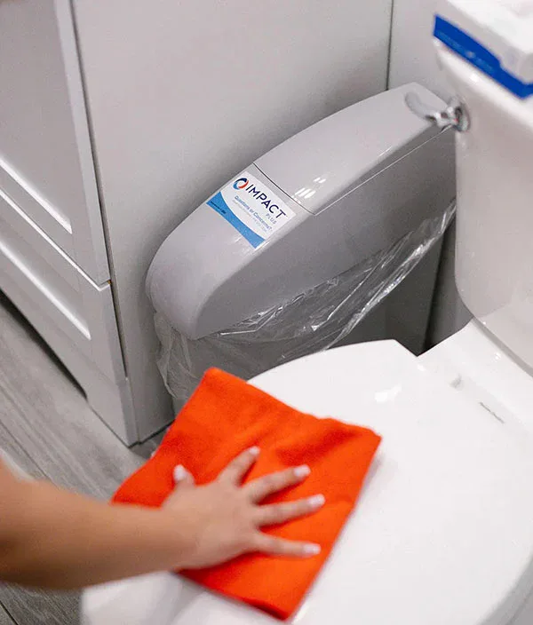 A person wipes a white toilet seat with an orange cloth. A toilet seat-cover dispenser labeled IMPACT is attached beside the toilet, reflecting the standards of impact commercial cleaning services. The bathroom has light-colored flooring and cabinets.