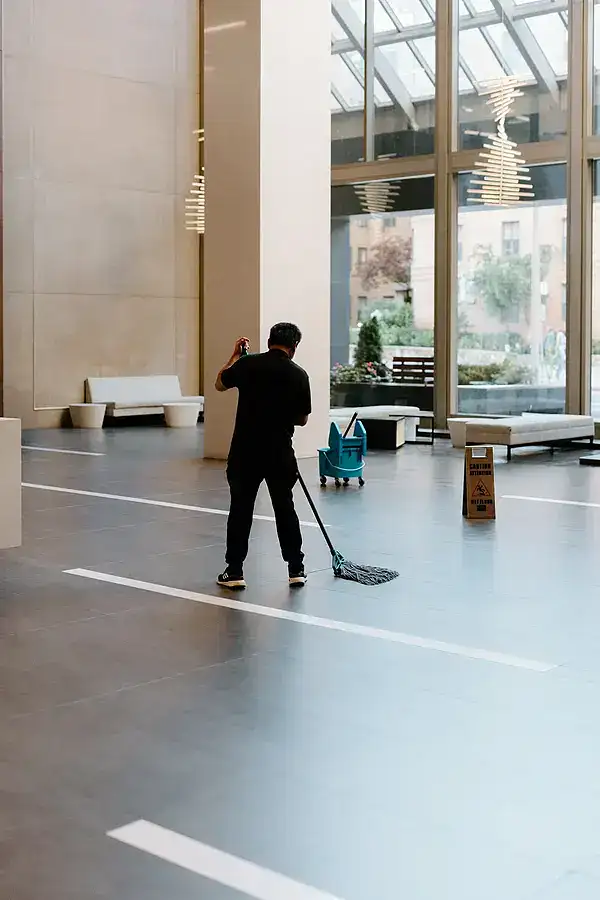 A person mops the floor in a spacious, modern building with large windows. Nearby are a blue cleaning bucket and a caution wet floor sign—showcasing the quality of impact commercial cleaning services. Benches and trees are visible through the windows.