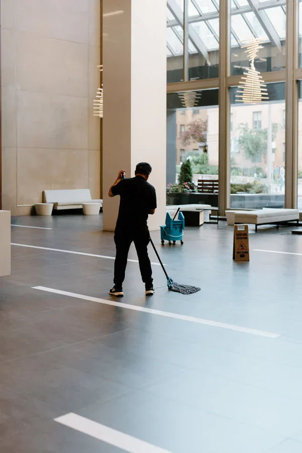 A person mops the floor in a spacious, modern building lobby with large windows, a blue mop bucket, and a caution wet floor sign nearby.