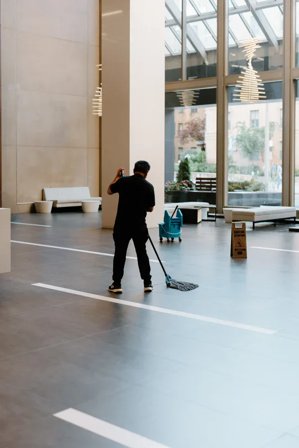 A person mops the floor in a spacious, modern lobby with large windows, benches, a blue cleaning cart, and a caution wet floor sign nearby.