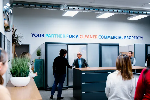 Several people are gathered in a modern office space. Two individuals near the center are shaking hands in front of a wall sign that reads, YOUR PARTNER FOR A CLEANER COMMERCIAL PROPERTY!, showcasing the impact commercial cleaning services in Canada.