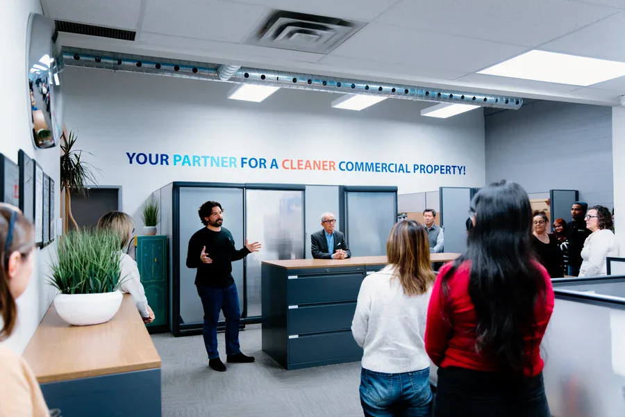 A group of people gathers in a modern office as a man speaks to them. Behind him, a wall sign reads, Your partner for a cleaner commercial property! Several people listen attentively.