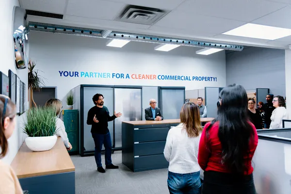 A group of people gathers in a modern office space while a man speaks to them. The wall behind him displays the slogan Your partner for a cleaner commercial property! promoting impact commercial cleaning services in Canada.