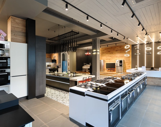 A spacious, modern kitchen showroom displays various kitchen appliances, including ovens, stoves, and refrigerators, with sleek countertops, wood accents, and stylish lighting fixtures.