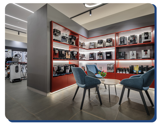 Modern showroom with shelves displaying a variety of coffee machines, two blue chairs, and a small round table with a flower centerpiece on a tiled floor. The space is brightly lit and neatly organized thanks to impact commercial cleaning services.
