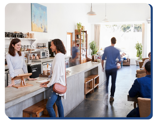A bright, modern coffee shop with people inside. A woman behind the counter smiles at a customer, while others chat at tables. Large windows fill the space with light, reflecting the cleanliness maintained by impact commercial cleaning services.