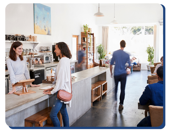 A bright, modern coffee shop with people chatting at the counter, a person walking toward the seating area, and others sitting at tables near large windows letting in natural light.