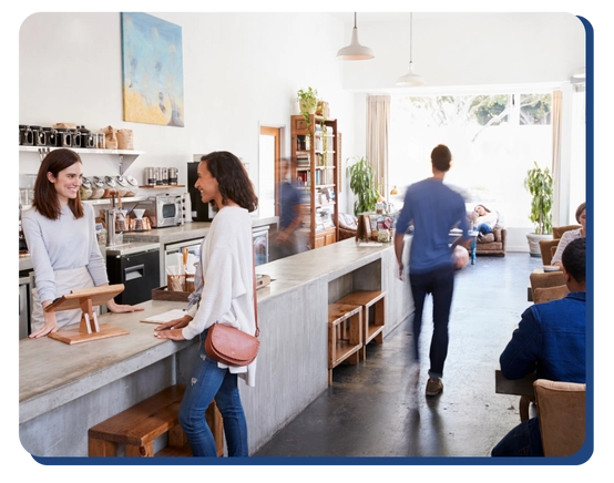 A bright, modern café with people chatting at the counter and others sitting at tables. Large windows let in sunlight, while shelves with plants and decor add a cozy touch—perfect for impact commercial cleaning services in Canada.