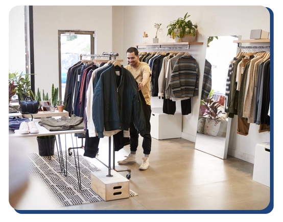 A man browses clothing on racks in a stylish, well-lit boutique with plants, shelves, and a large mirror. The impact of commercial cleaning services in Canada keeps the casual and neatly arranged clothes on display throughout the space.