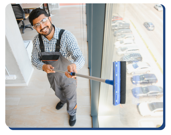 A smiling man in work overalls and glasses cleans a large window with a squeegee in a modern building, showcasing the professionalism of impact commercial cleaning services, with a view of parked cars and a road outside.