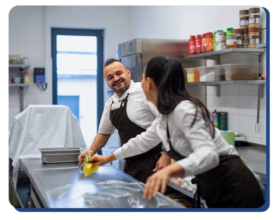 Two people in aprons smile as they clean a stainless steel counter in a commercial kitchen. Shelves with containers and spices are visible in the background.