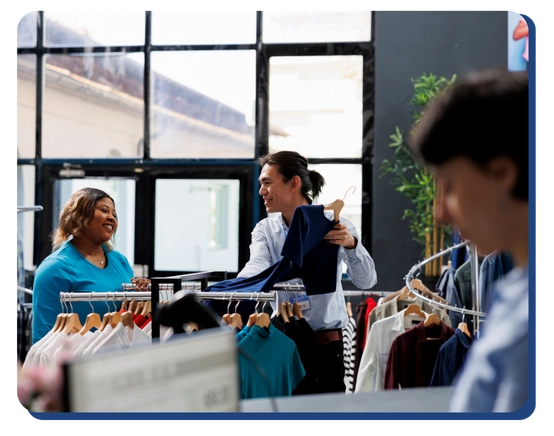 Two people smile and talk while browsing clothes on racks in a bright, modern clothing store. Another person is blurred in the foreground. Sunlight comes through large windows in the background.