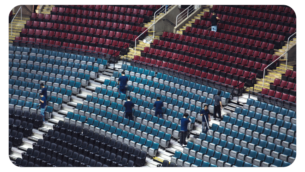Workers in blue shirts are spaced out, cleaning or inspecting rows of empty seats in a large, multi-tiered stadium with maroon and teal seats.