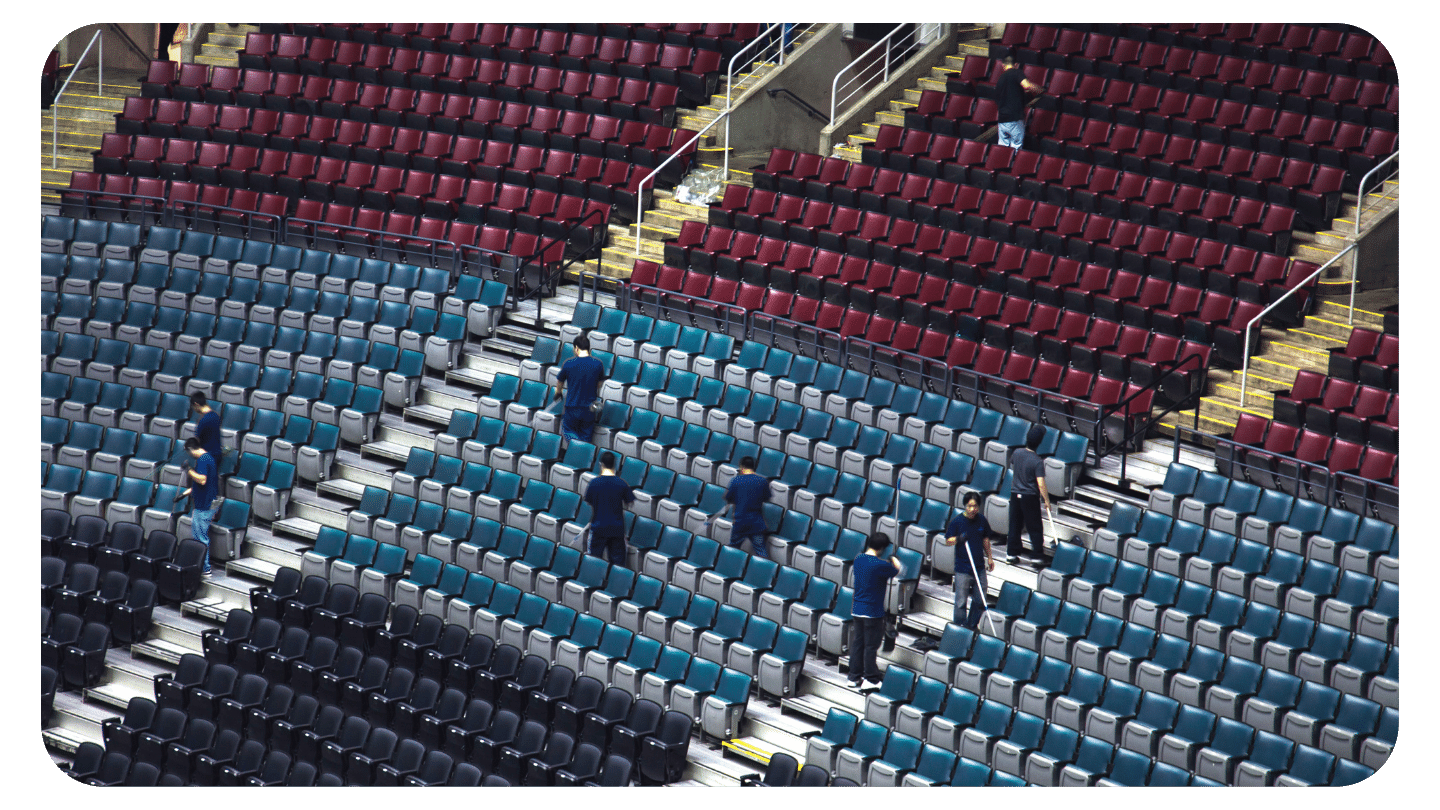Workers in blue shirts are spaced out, cleaning or inspecting rows of empty seats in a large, multi-tiered stadium with maroon and teal seats.