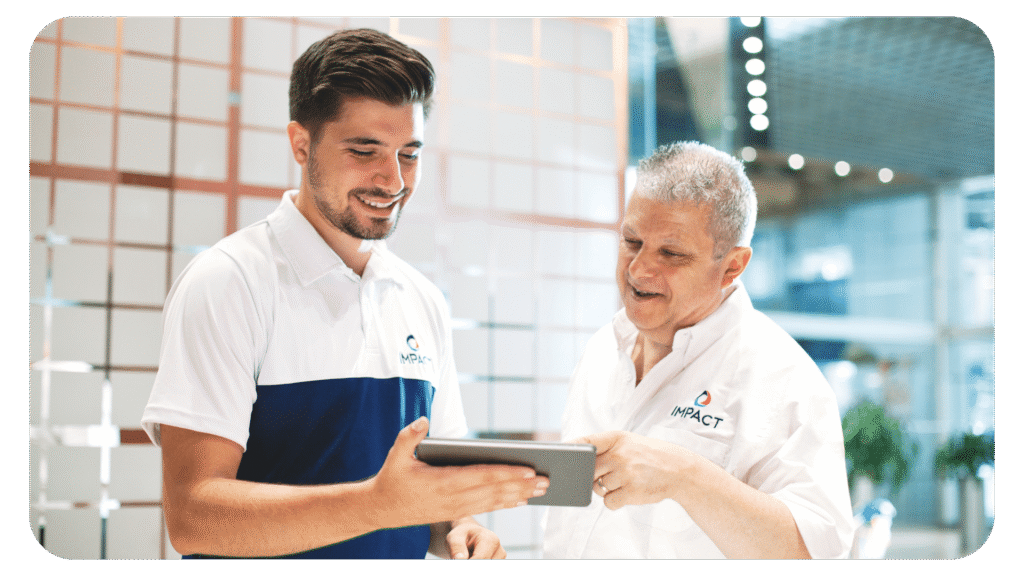 Two men wearing “IMPACT” branded shirts smile while looking at a tablet together in a modern office setting with glass walls and decorative panels.