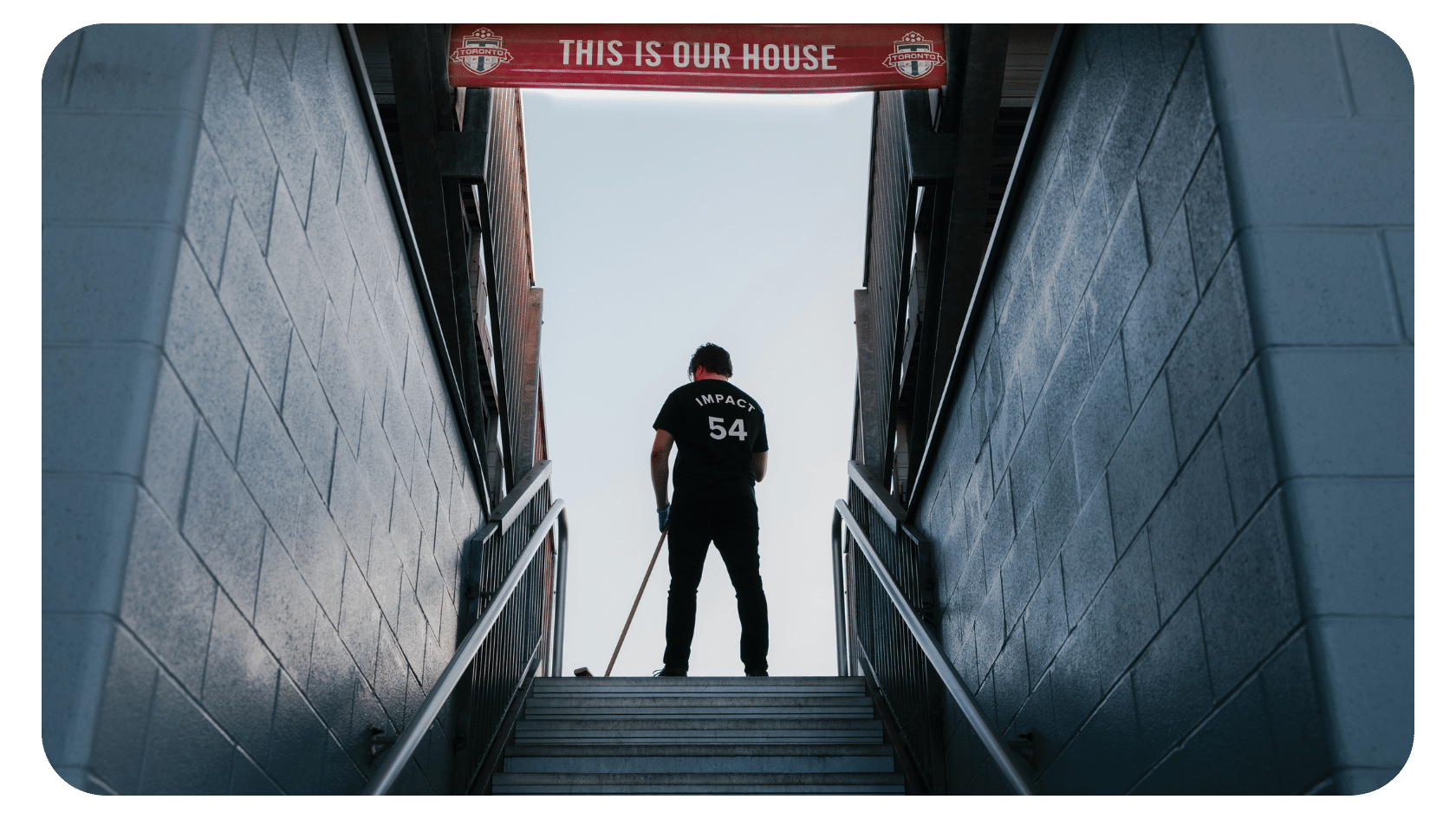 A person in a “IMPACT 54” shirt stands at the top of outdoor stairs, facing away, under a red banner that reads THIS IS OUR HOUSE, with sunlight in the background.