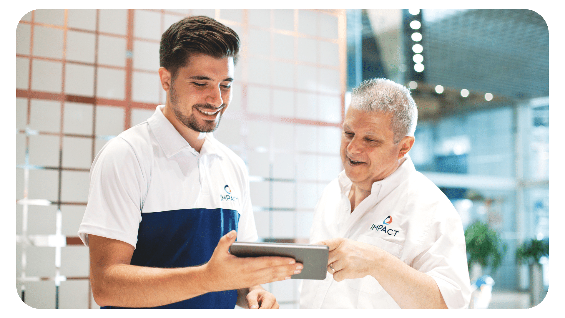 Two men wearing “IMPACT” branded shirts smile while looking at a tablet together in a modern office setting with glass walls and decorative panels.
