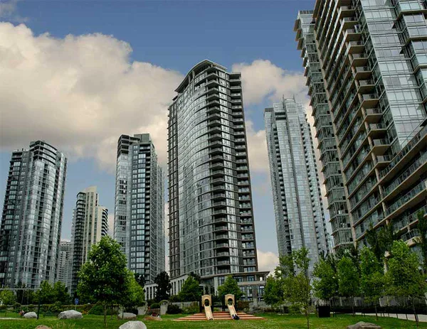 Modern high-rise apartment buildings surround a small green park with trees, grass, rocks, and a playground under a partly cloudy sky, all maintained by impact commercial cleaning services.