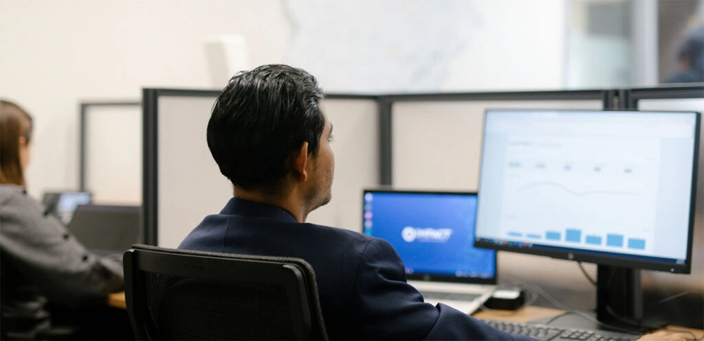 Two people work at office desks with computers. The man in the foreground views a monitor displaying charts and graphs, while a woman types at another desk in the background. The workspace is sectioned by dividers.