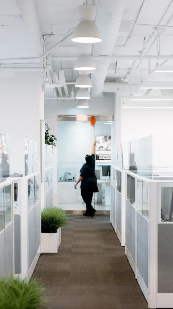 A person from Impact Commercial Cleaning Services cleans a light fixture on the ceiling in a modern, bright office hallway with white walls, glass partitions, and potted plants along the corridor.