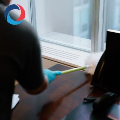 A person wearing blue gloves dusts a wooden desk near a window with a white duster. A black telephone and monitor are on the desk, and a logo is visible in the top left corner.