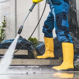 A person wearing yellow rubber boots and blue work pants uses a pressure washer to clean outdoor stone steps, showcasing the thorough results of impact commercial cleaning services.