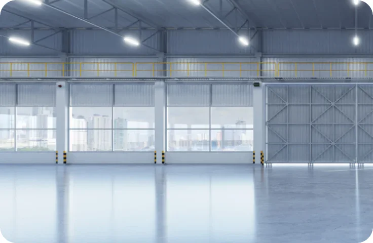 Spacious, empty industrial warehouse with large windows, high ceilings, and polished concrete floors; city buildings visible outside through the windows.